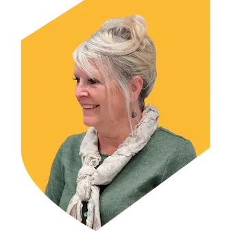 Woman with blonde hair in a green top with a white scarf sitting with a yellow background in the shape of a shield.